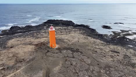 Drohnenaufnahmen-Fangen-Die-Schönheit-Eines-Farbenfrohen-Leuchtturms-An-Der-Küste-Islands-Ein,-Der-Auf-Einem-Schwarzen-Sandstrand-Inmitten-Einer-Dramatischen-Landschaft-Aus-Felsen-Und-Meer-Thront