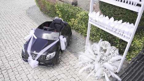 miniature-Mercedes-Benz-toy-car-decorated-with-white-ribbons,-surrounded-by-wedding-accessories-on-a-cobblestone-path