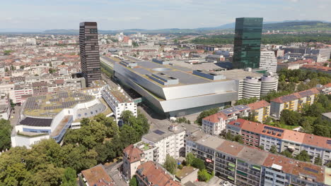 Edificio-De-La-Feria-De-Basilea-Con-Paisaje-Urbano-Y-Camión-En-Movimiento-En-Un-Día-Soleado,-Vista-Aérea