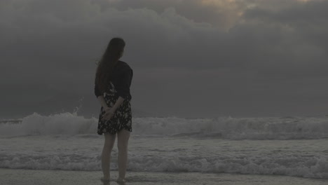 Pondering-young-female-wearing-glasses-looking-out-across-stormy-overcast-ocean-waves,-rear-view