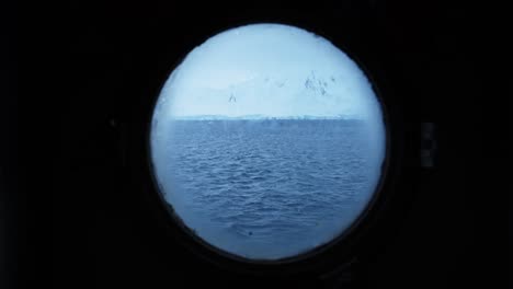 Vista-De-Ojo-De-Buey-En-El-Barco-De-La-Antártida-Con-La-Península-Antártica,-El-Océano,-El-Agua-Del-Mar-Y-El-Hielo-Helado-Cubierto-De-Tierra-Nevada-Cubierta-De-Nieve