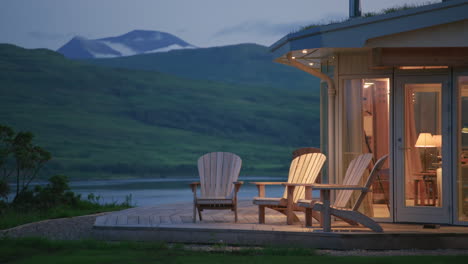 Beleuchtetes-Haus-Mit-Adirondack-Stühlen-Auf-Holzterrasse-Mit-Blick-Auf-Den-Malerischen-See