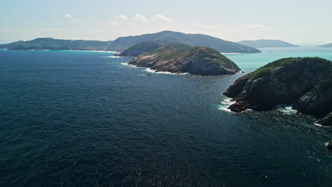 Drone-soars-over-Arraial-do-Cabo's-coastal-waters,-showcasing-stunning-rocky-shorelines-with-majestic-mountains-in-the-background,-capturing-nature’s-raw-beauty-from-above