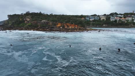 Aerial-Pan-of-Surfers-in-Lineup-on-Ocean-Waves-Cloudy-Summers-Day-Rocky-Coastline-Shelly-Headland,-Sydney,-Australia