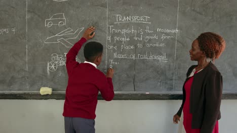 Boy-And-His-Teacher-Writing-On-Blackboard-In-African-School---Medium-Shot