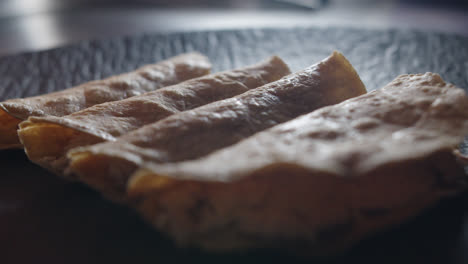 Hermosa-Toma-En-Primer-Plano-Y-En-Cámara-Lenta-De-Un-Grupo-De-4-Enchiladas-De-Pollo-Colocadas-En-Un-Plato-Negro-Y-Retroiluminadas-En-Una-Cocina-Industrial-De-Un-Restaurante.