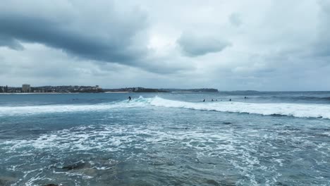 Toma-Aérea-Baja-Sobre-La-Costa-Rocosa-De-Sydney,-Volando-Sobre-El-Agua-Y-Levantándose-Para-Revelar-A-Un-Surfista-Despegando-Sobre-Las-Olas-Del-Océano-En-Manly,-Australia