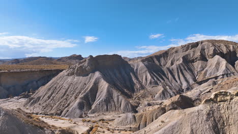 Flight-curves-right-exploring-the-iconic-rugged-landscape-of-Llano-del-Duque-and-Rambla-del-Infierno-with-grey-eroded-hillsides,-and-tufts-of-brown-grasses-under-intense-heat-and-sunshine,-Tabernas