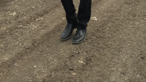 A-man-dressed-in-a-white-t-shirt-and-dark-pants-strolls-along-a-dirt-road-surrounded-by-expansive-green-fields