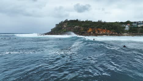 Zeitlupen-Luftaufnahme-Von-Meereswellen,-Die-Ans-Ufer-Schlagen,-Mit-Surfern,-Die-In-Einer-Reihe-Warten,-Offshore-Windsurfen-An-Der-Felsigen-Küste-In-Sydney,-Australien