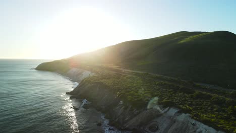 Drone-pullback-above-grassy-green-hills-sloping-down-to-ocean-with-sunlight-peaking-over-hills