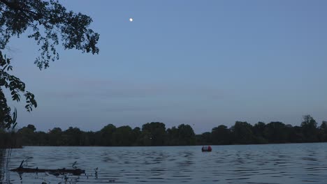 Toma-Panorámica-De-La-Luna-Al-Anochecer-Sobre-Un-Lago-Tranquilo-En-Un-Parque-Con-árboles.