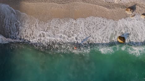 Overhead-aerial-view-of-a-tourists-bathing-in-seawater,-Sea-waves-touching-shore,-Albanian-Riviera-secluded-Filikuri-Beach