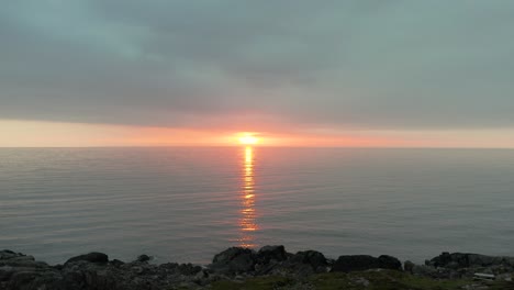 Aerial-pullback-over-serene-water-retreating-from-orange-red-glow-of-light,-Farstadsanden-beach-at-sunset-in-Norway