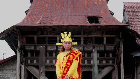 Beautiful-young-woman-in-a-traditional-yellow-dress-in-front-of-an-old-house-in-Iompat-Batu,-Desa-Bawomataluo,-Bawömataluo