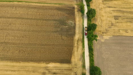 Vista-Aérea-De-Un-Campo-De-Trigo-Cosechado-Con-Un-Tractor-Circulando-Por-Un-Camino-Rural,-Bordeado-De-árboles.