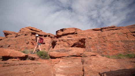 Young-Woman-With-Backpack-Walking-on-Red-Sandstone-Hill-in-Desert-Landscape-on-Hot-Sunny-Day