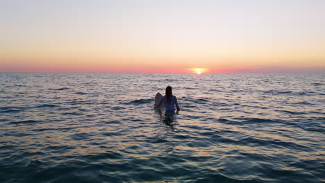 Woman-sitting-on-surfboard-in-the-ocean-at-sunset