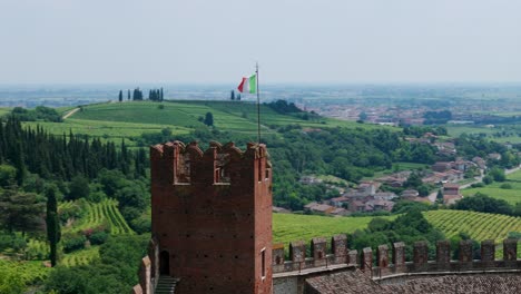 Luftaufnahme-Des-Schlossturms-Von-Soave-Mit-Der-Italienischen-Flagge-Mit-Blick-Auf-Die-üppige-Grüne-Landschaft