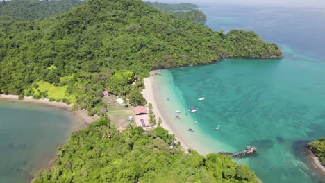 Aerial-view-of-the-tropical-Coiba-Island-beach-in-Panama,-featuring-lush-greenery,-clear-turquoise-waters-and-boats-along-the-shore