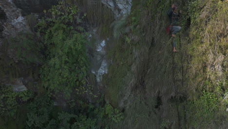 Drone-shot-of-a-mad-honey-hunter-climbing-down-a-rope-on-bamboo-stairs-at-a-cliff-in-Lamjung,-Nepal,-after-mad-honey-extraction-Highlights-the-adventurous-and-risky-agricultural-practice