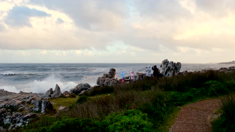 Two-adults-with-young-two-kids-watch-sea-waves-crash-spectacularly-against-rocks