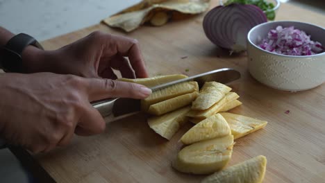 Weite-Aufnahme-Einer-Reifen-Gelben-Kochbanane,-Die-In-Kleine-Stücke-Geschnitten-Wird,-Und-Spezieller-Zutaten-Zum-Kochen-Einer-Mahlzeit,-Zwei-Dosen-Bohnen,-Reis,-Kochbanane,-Avocado,-Rote-Zwiebeln-Und-Koriander