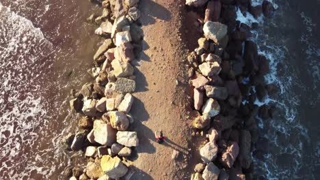 Lonely-man-walking-rocky-patch-surrounded-by-ocean-sea-water,-top-down-aerial-view