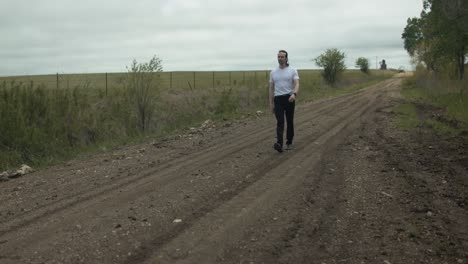 A-man-dressed-in-a-white-t-shirt-and-dark-pants-strolls-along-a-country,-dirt-road-surrounded-by-expansive-green-fields