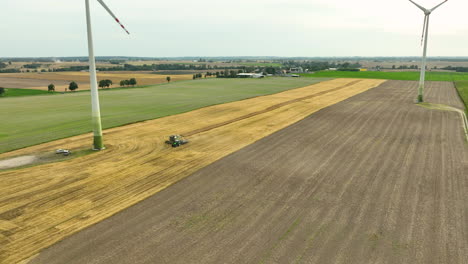 Vista-Aérea-De-Turbinas-Eólicas-En-Un-Vasto-Paisaje-Agrícola,-Con-Un-Tractor-Trabajando-En-Un-Campo-De-Trigo.