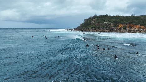 Low-aerial-angle-of-Surfers-taking-off-on-Ocean-Waves-on-Cloudy-Day-infront-of-Rocky-Coastline-in-Sydney-Australia-Slowmo-Video