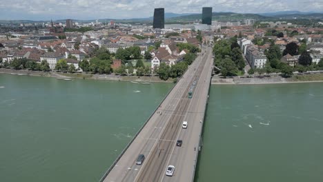 Vídeo-De-Dron-En-4K-Del-Tren-Que-Cruza-El-Puente-Wettsteinbrücke-Sobre-El-Río-Rin-En-Dirección-A-Los-Modernos-Rascacielos-De-Basilea,-Suiza