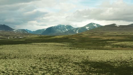 Plataforma-Aérea-Sobre-Pastizales-De-Color-Verde-Claro-Y-Oscuro-Del-Parque-Nacional-De-Rondane,-Condado-De-Innlandet,-Noruega,-Con-Picos-De-Montaña-A-Lo-Lejos