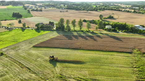 Vista-Aérea-De-Un-Tractor-Cosechando-En-Exuberantes-Campos-Verdes,-Con-Patrones-Contrastantes-De-Marrón-Y-Verde-Y-Un-Hermoso-Paisaje-Rural-En-El-Fondo.