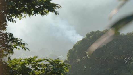 Heavy-smoke-rising-from-small-village-in-jungle-landscape,-handheld