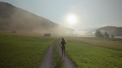 Woman-running-on-a-dirt-path-through-misty-meadow-at-sunrise,-slow-motion