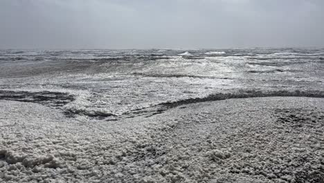 Zitternder-Meeresschaum-Weht-Nach-Einem-Sturm-An-Der-Küste-Auf-Den-Strand,-Monochrom