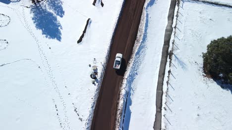 Aerial-tracking-follows-white-truck-driving-along-rural-dirt-road-with-snow-covered-fields