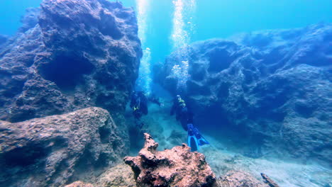 Two-scuba-divers-swimming-through-a-narrow-passage-between-large-rocks-in-a-clear-blue-ocean