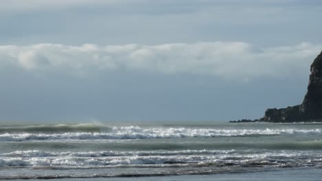 Surfers-are-just-visible-close-to-rocky-headland-on-a-blustery-winter's-day---Whitewash-Head,-Scarborough,-New-Zealand