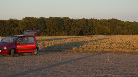 Small-red-car,-unidentified-brand-parked-near-field-and-dense-forest,-camping