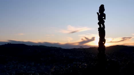 Scenic-view-of-glowing-sunset-behind-cultural-Maori-pou-whenua-carved-statue-overlooking-the-capital-city-of-Wellington,-New-Zealand-Aotearoa