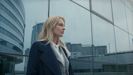 A-businesswoman-with-blonde-hair-stands-outside-a-glass-building-in-London,-her-confident-stance-and-professional-attire-making-a-striking-impression-against-the-sleek,-modern-architecture