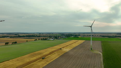 Aerial-view-of-wind-turbines-in-an-expansive-agricultural-area,-with-a-tractor-working-on-a-harvested-field