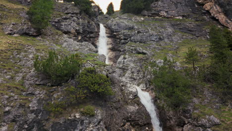 Aerial-view-of-twin-waterfalls-cascading-over-rocky-cliffs-in-the-Dolomite-mountains,-surrounded-by-lush-greenery