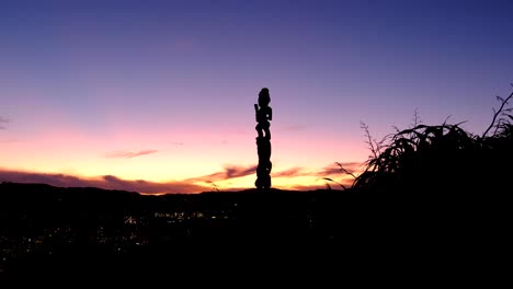 Estatua-De-Madera-Tallada-De-Un-Maorí-Pou-Whenua-En-Silueta-Y-Plantas-De-Lino-Contra-Un-Impresionante-Cielo-Púrpura-Al-Atardecer-En-Wellington,-Nueva-Zelanda,-Aotearoa