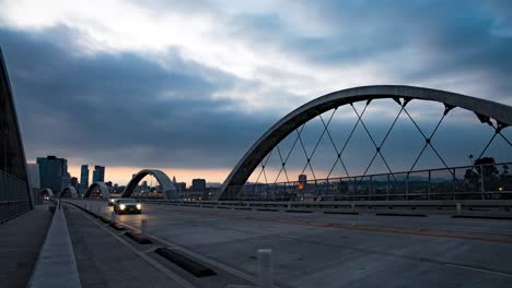 Un-Lapso-De-Tiempo-De-La-Cinta-De-Luz-Del-Puente-De-La-Calle-6-Durante-La-Puesta-De-Sol-En-Los-Ángeles,-California,-Con-Automóviles-Circulando-En-El-Primer-Plano-Del-Horizonte-De-La-Ciudad.