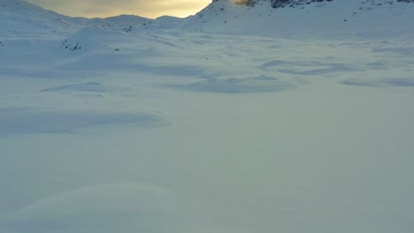 Drohne-Schwenkt-Nach-Oben-über-Schneehügel,-Während-Die-Sonnenstrahlen-Der-Untergehenden-Sonne-Wolken-Und-Landschaft-Gelb-Erhellen,-Haukelifjell-Bergpass-In-Vinje-Telemark,-Norwegen-Bei-Sonnenuntergang