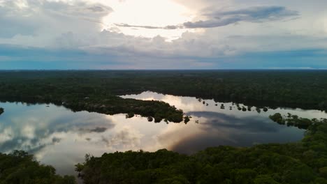 Erleben-Sie-Eine-Faszinierende-Luftaufnahme,-Während-Eine-Drohne-Nach-Links-Gleitet-Und-Die-üppigen-Baumkronen-Des-Amazonas-Regenwalds-Und-Die-Ruhige-Spiegelung-Des-Himmels-Und-Der-Sonne-Auf-Dem-Amazonas-Fluss-Darunter-Einfängt