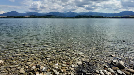 Crystal-clear-lake-water-with-rocky-beach-and-mountains